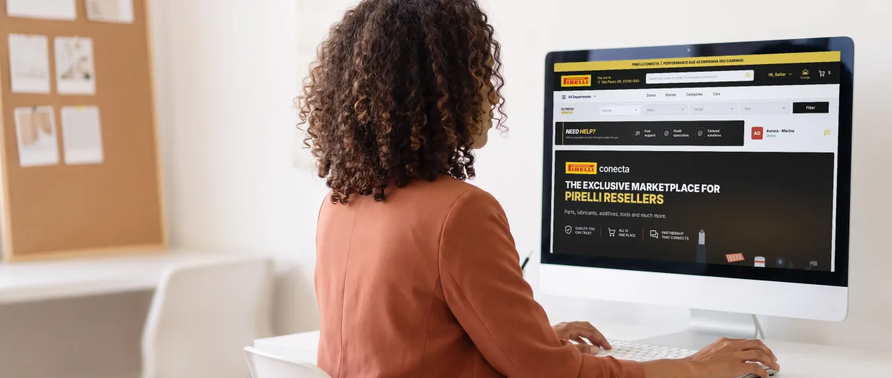 CMO with curly hair and a terracotta blazer, seated at a desk facing the monitor; the screen shows the Pirelli Conecta home page, the marketplace exclusive to resellers.