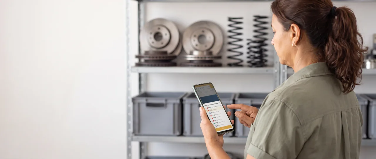 Buyer in a work shirt, in an auto parts shop, looks at the phone in her hand; the screen shows the store picker on the Fortbras portal.