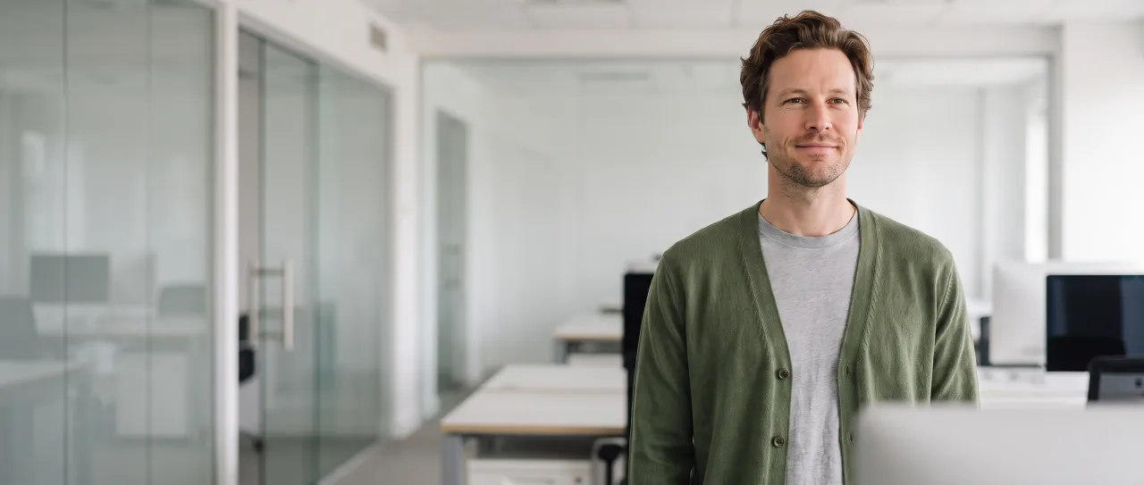 Diretor de tecnologia sorri de leve, tranquilo, num escritório aberto e claro, com mesas de trabalho e divisória de vidro desfocadas ao fundo, de dia.