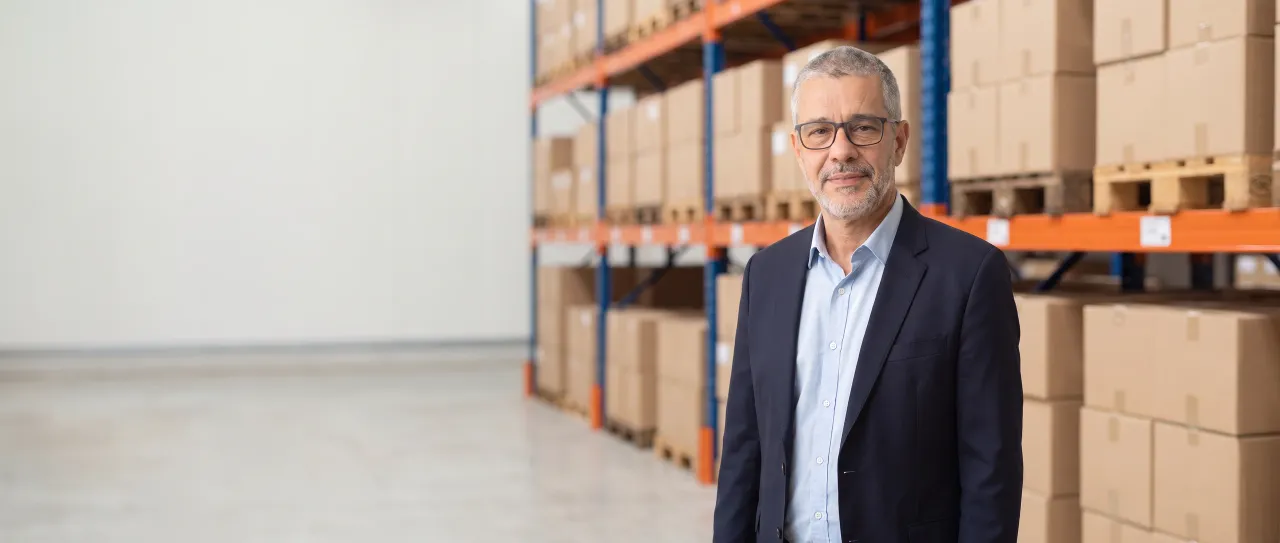 Diretor de uma distribuidora, de blazer azul-marinho e óculos, sorri tranquilo no corredor claro do centro de distribuição, com porta-paletes desfocados.