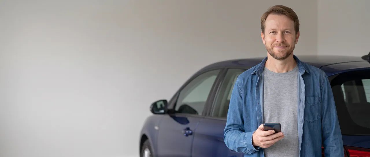 Dono de um carro sorri satisfeito na garagem clara de casa, com o celular na mão e o veículo visto de lado e desfocado ao fundo, à luz do dia.