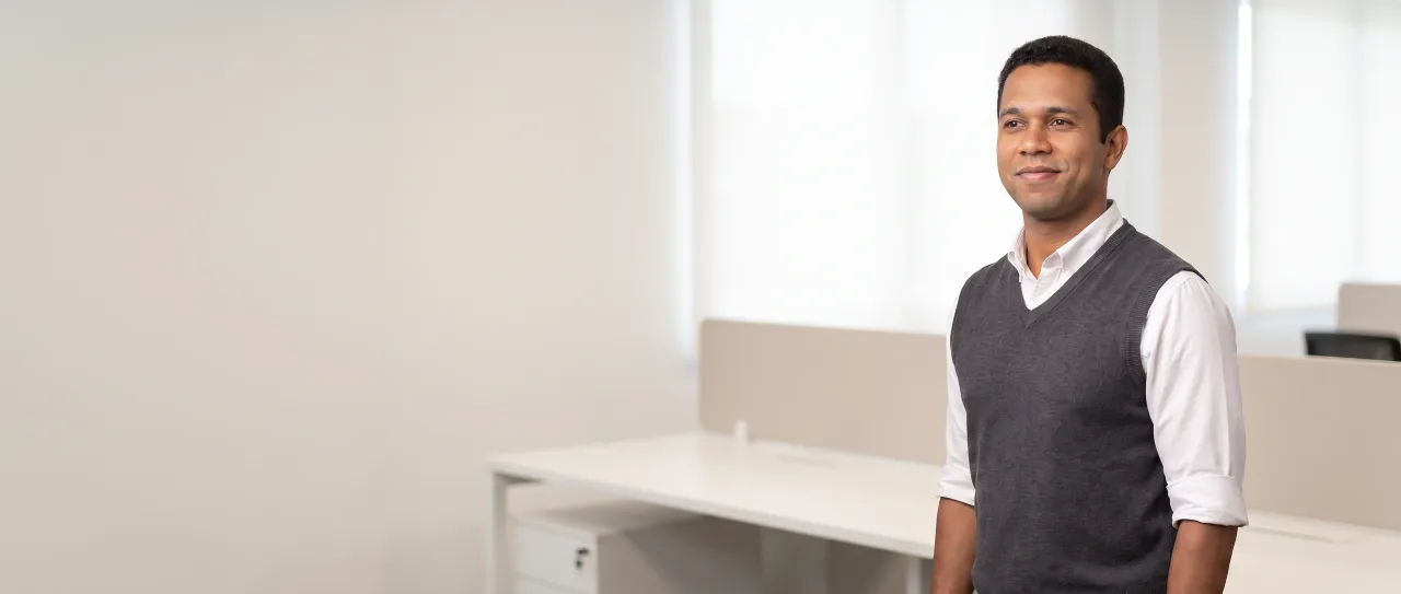 Representante comercial sorri, tranquilo, de pé numa sala comercial clara, com mesas de trabalho e janelas de luz difusa desfocadas ao fundo, de dia.