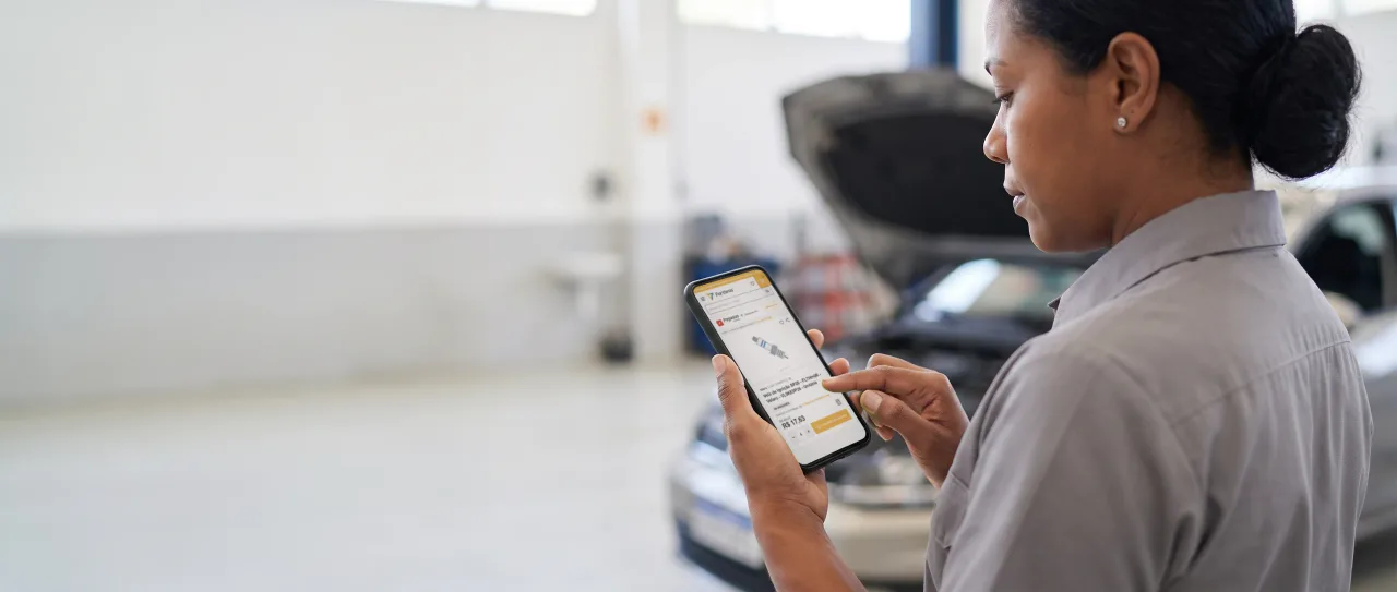 Técnica de oficina, de uniforme cinza, olha o celular na mão, com um carro de capô aberto ao fundo; a tela mostra uma peça com o estoque da filial no portal da Fortbras.