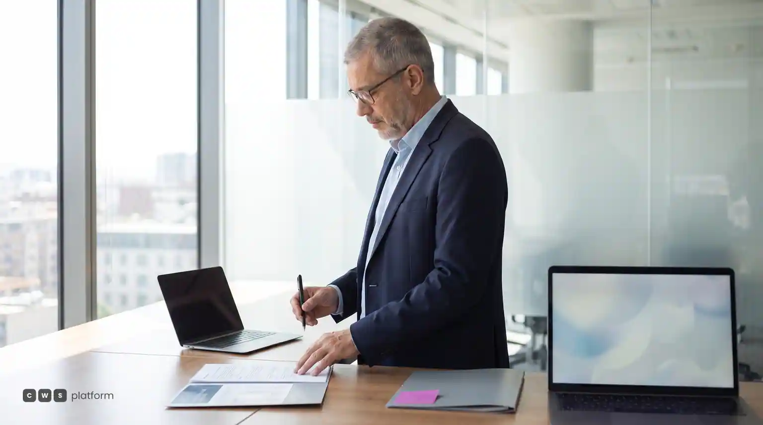 Diretor comercial de óculos revisando e assinando papéis numa mesa com dois notebooks.